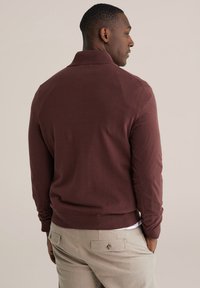 Pull-over bordeaux avec un col côtelé, des manches longues et un tissu texturé, associé à un pantalon beige clair avec des poches arrière.