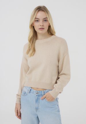 Maglione cropped beige con colletto rotondo e polsini a coste, abbinato a jeans azzurri, creando un look casual e rilassato.