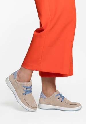 Bež antilop tenisice s plavim pertlama i detaljem na peti, nošene uz svijetlosmeđe kratke hlače na svijetloj pozadini.