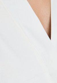 Tessuto bianco con una superficie liscia, caratterizzato da un design a V e da un sottile dettaglio di cucitura lungo il colletto. Vista ravvicinata.