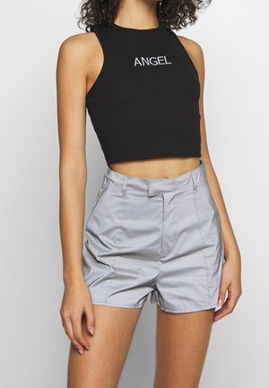 Jong persoon draagt een zwart mouwloos cropped top met tekst "ANGEL" en lichtgrijze shorts met hoge taille, staand tegen een effen achtergrond.