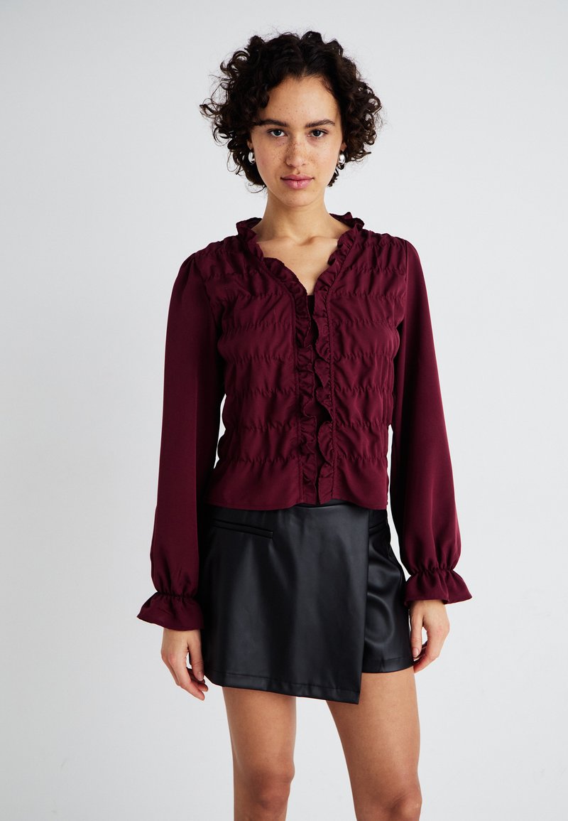 Μπουφόνα μπορντό με ρυτίδες και μακριά μανίκια, συνδυασμένο με μαύρη ψεύτικη δερμάτινη φούστα με ασύμμετρη κοπή και σχέδιο τσέπης.