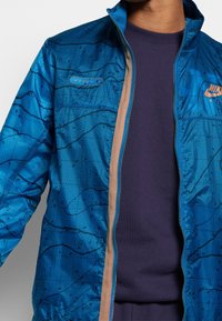 Sininen kevyt takki vetoketjulla edessä, jossa on geometrinen kuvio ja Nike-brändäys rinnassa. Sisäpuolella on kontrastivärinen tan-vuori.