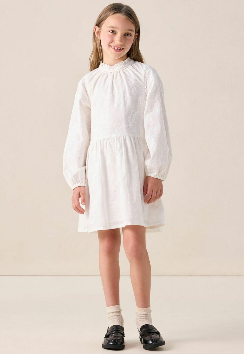 Jeune fille souriante, portant une robe blanche à manches longues avec des motifs floraux subtils, des chaussettes crème et des chaussures noires brillantes, se tenant sur un fond clair.