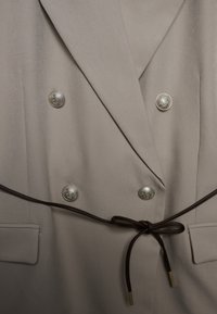 Beiger zweireihiger Blazer mit silbernen Knöpfen, braunem Gürtelakzent und zwei Fronttaschen; glatter, maßgeschneiderter Stoff mit strukturierter Gestaltung.