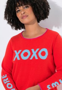 Kobieta z kręconymi włosami w czerwonym swetrze z niebieskim napisem "XOXO" na piersi oraz dodatkowymi niebieskimi napisami na rękawach.