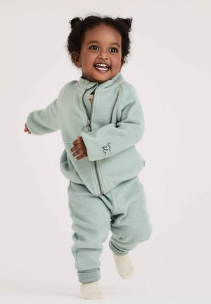 Lächelndes Kleinkind, das in einer hellgrünen Fleecejacke und -hose mit beigen Socken vor einem schlichten Hintergrund läuft.