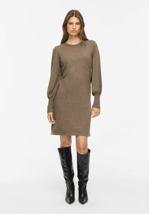 Femme portant une robe en maille marron à manches longues et des bottes en cuir noires montant jusqu'aux genoux, debout devant un fond blanc uni.