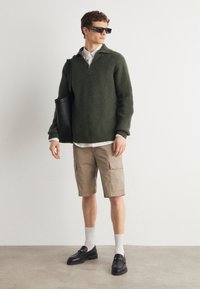 Pullover verde lavorato a maglia con colletto zip, pantaloni cargo beige, mocassini neri e borsa a tracolla nera. Il modello indossa occhiali da sole rettangolari.