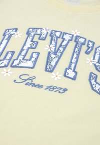 Ανοιχτό κίτρινο μπλουζάκι με μπλε γράμματα "LEVI’S" σε στυλ δαντέλας και λευκά μαργαρίτες. Φέρει τη φράση "Since 1873" σε καλλιγραφική γραφή. Από βαμβακερό υλικό, με κοντά μανίκια.