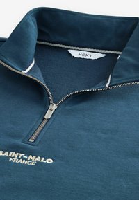 Felpa blu con zip, caratterizzata da una texture morbida, colletto alto, zip argentata e ricamo "SAINT-MALO FRANCIA" in bianco.