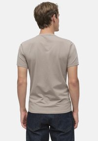 Ung man med kort brunt hår som bär en enkel beige t-shirt med korta ärmar och mörkblå jeans, står med ryggen mot kameran.