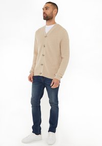 Beige gestrickte Strickjacke mit V-Ausschnitt, Knopfleiste und strukturiertem Muster, kombiniert mit dunkelblauen Jeans und weißen Sneakers.