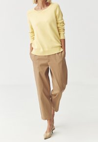Jasnożółty sweter z dzianiny o szerokim dekolcie i ribbowym wykończeniu, w połączeniu z beżowymi spodniami o luźnym kroju. Buty to jasnobeżowe, szpiczaste szpilki.