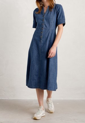 Denim-mekko, jonka pituus on keskipitkä, lyhyet hihat, napitettu edestä, V-kaula-aukko ja paneelisaumat. Yhdistetty vaaleisiin urheilukenkiin.