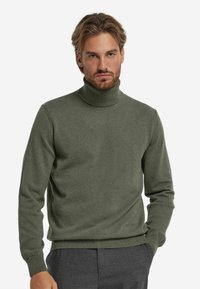 Grüner Rollkragenpullover mit gerippten Bündchen und Saum, weicher Textur, tailliertem Design, getragen über maßgeschneiderten grauen Hosen.