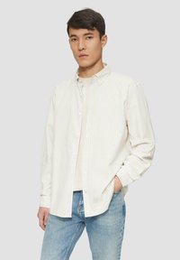 Beige overhemd met lichte verticale strepen, lange mouwen, een kraag en een relaxte pasvorm, gedragen over een crèmekleurig shirt. Lichte spijkerbroek.