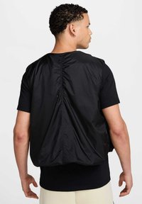 Sort nylon vest med cinch på ryggen, minimalistisk design og uden ærmer. Letvægtsstof med blød tekstur.