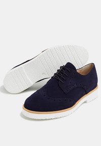 Chaussure brogue en suède bleu marine avec garniture beige, semelle en caoutchouc blanche, lacets noirs et détails décoratifs perforés.