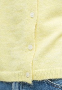 Hele kollane kardigan pehmest, tekstuurse kudumiga, millel on neli valget nööpi ees ning soonikuga ääreosa, kombineeritud teksapükste vööosaga.