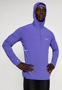 Lila sportig tröja med halvdragkedja fram, huva och reflexdetalj. Har långa ärmar och elastiskt material. Logotyp på bröstet.
