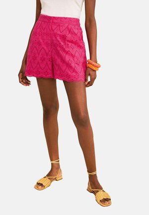 Woman wearing bright pink patterned shorts, white top, orange bracelets, and beige woven sandals with ankle straps, standing against a white background.