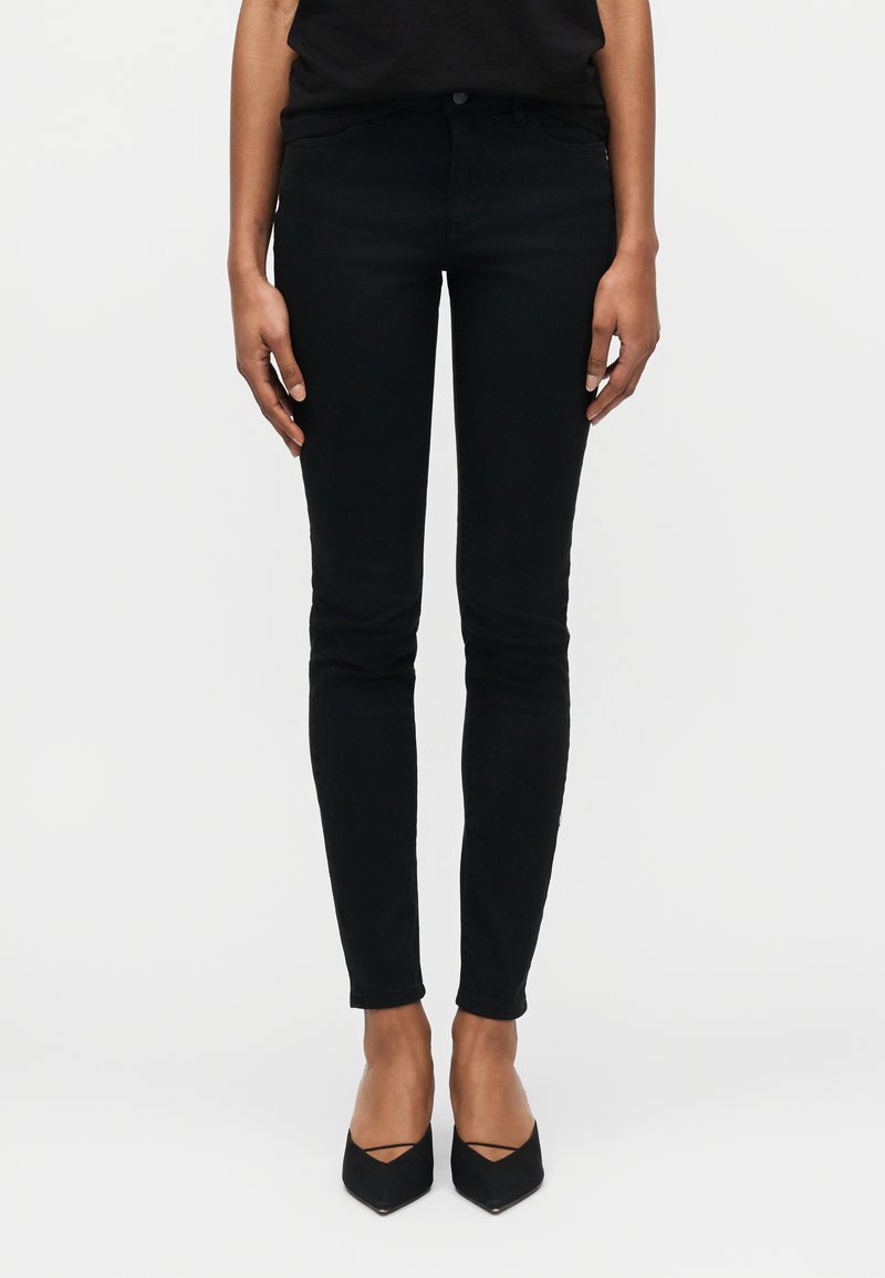 Γυναίκα με μαύρα στενά τζιν και μαύρες μυτερές γόβες, στέκεται με τα χέρια χαλαρά στο πλάι μπροστά σε απλό φόντο.