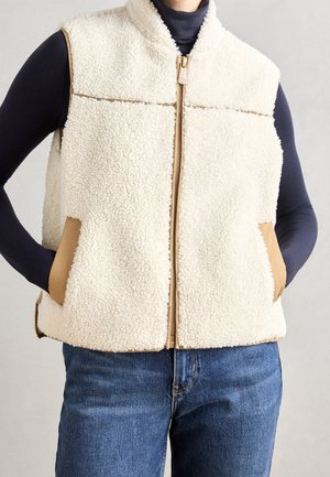 Personne portant un gilet sans manches en polaire crème zippé sur un haut noir à manches longues et un jean bleu, les mains dans les poches du gilet.