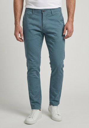 Homme portant un pantalon slim bleu sarcelle et des baskets blanches, debout devant un fond uni.