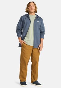 Mandens afslappede outfit med en marineblå zip-hoodie, lys grøn t-shirt og brune bukser, samt blå sneakers. Glatte teksturer, afslappet pasform.