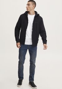 Svart zip-up hettegenser med snøring, brukt over en hvit t-skjorte, sammen med blå jeans og svarte joggesko. Mykt materiale, uformell passform.