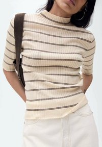 Lichtbeige ribbelige top met korte mouwen, verticale bruine strepen, hoge halslijn en cropped zoom, gedragen met een schoudertas.