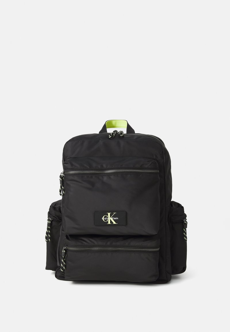 Black fabric backpack with green accent, multiple zippered pockets, and logo patch. Features textured straps and contrasting drawstring.