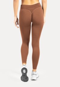 Braune Leggings mit hoher Taille, glatter Textur, figurbetontem Design und Konturnaht-Details, kombiniert mit weißen Sportschuhen.