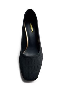 Zapato de tela negro con punta cuadrada, textura acanalada y un interior suave. Detalle de marca dorado visible en el interior del zapato.