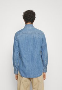 Ljusblå denimskjorta med långa ärmar, knäppningar vid ärmsluten, en spetsig krage och en rundad nederkant, som visar en lätt urblekt textur.