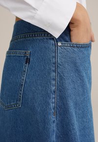 Džínová sukně v tmavě modré barvě, s postranním švem, zadní kapsou a viditelným zapínáním na knoflíky. Materiál má hladkou strukturu s mírným blednutím.