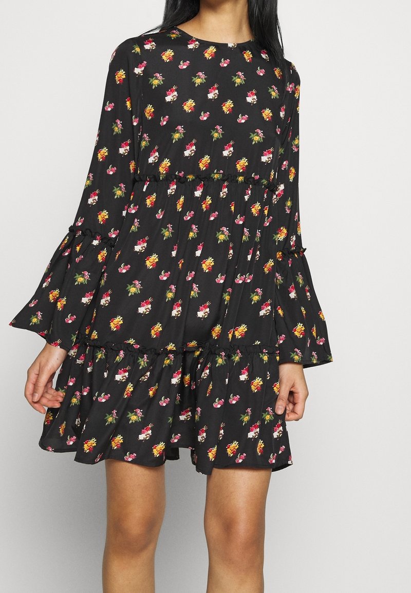 Svart klänning med blommönster i rosa, gult och vitt. Har långa klockärmar, en flerstegs nederdel och volangdetaljer vid sömmarna.