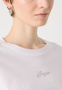 White cotton T-shirt with a scoop neckline and a silver rhinestone logo accent on the left side. Smooth texture, casual design.