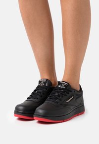 Chaussures de sport noires avec tige en cuir texturé, semelle en caoutchouc rouge et lacets noirs. Présentent un logo Reebok et des perforations pour la respirabilité.