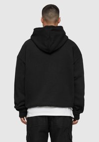 Felpa nera con cappuccio grande, vestibilità oversize e spalle scese. Polsini e orlo a coste, indossata sopra una maglietta bianca e pantaloni neri.