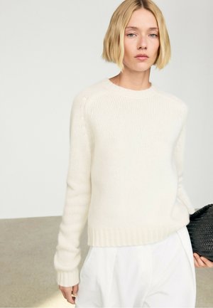 Vrouw met kort blond haar draagt een witte gebreide trui en witte broek, houdt een zwarte gestructureerde tas vast en staat tegen een effen achtergrond.