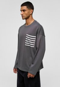 Donkergrijze sweatshirt met lange mouwen en een ronde hals, met een gestreepte patchzak aan de linkerkant. Losse pasvorm, zachte stofstructuur.