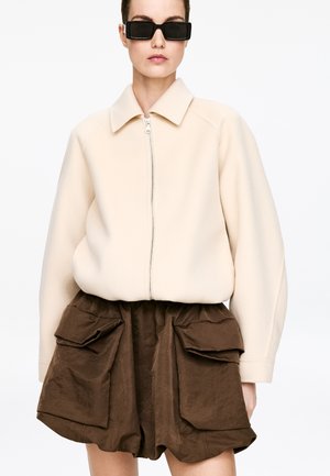 Kobieta w beżowej kurtce na suwak i brązowych szortach z wysokim stanem oraz dużymi przednimi kieszeniami z patkami, w czarnych prostokątnych okularach przeciwsłonecznych.