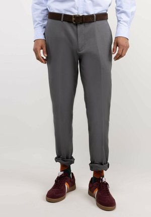 Hombre con pantalones grises remangados en los puños, un cinturón marrón, camisa azul clara, calcetines coloridos y zapatillas burdeos.