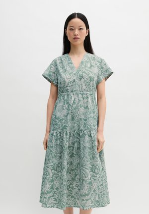 Kvinde stående iført en grøn og hvid blomsterprintet V-hals kjole med korte ærmer og flæset nederdel, mod en ensfarvet hvid baggrund.