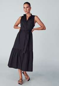 Abito nero senza maniche con collo a camicia, vita con cintura e orlo a strati, caratterizzato da uno spacco frontale e una trama liscia. Indossato con sandali neri.