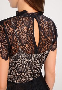 Svart spetsklänning med hög halsringning, korta ärmar och en nyckelhålsrygg. Spetsen har ett blommönster som avslöjar ett nakenfärgat underlager.