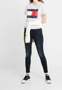 Grå bomulls t-shirt med "TOMMY JEANS" logotyp, svarta skinny jeans, vit stickad tröja knuten i midjan, och svarta sneakers med vita sulor.