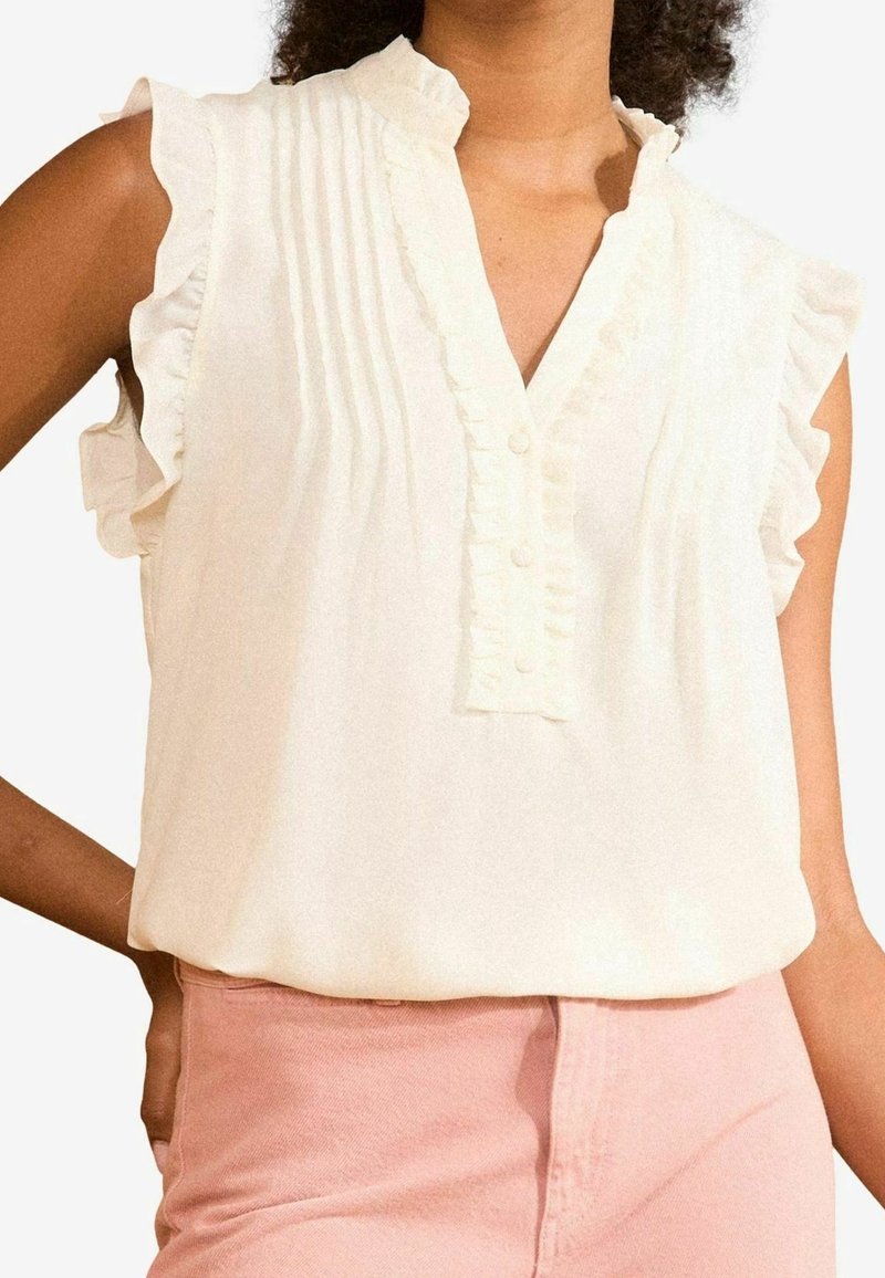 Vit ärmlös blus med volangdetaljer på axlarna, knappslå och vertikala veck. Bärs med ljusrosa byxor. Mjuk tygstruktur.